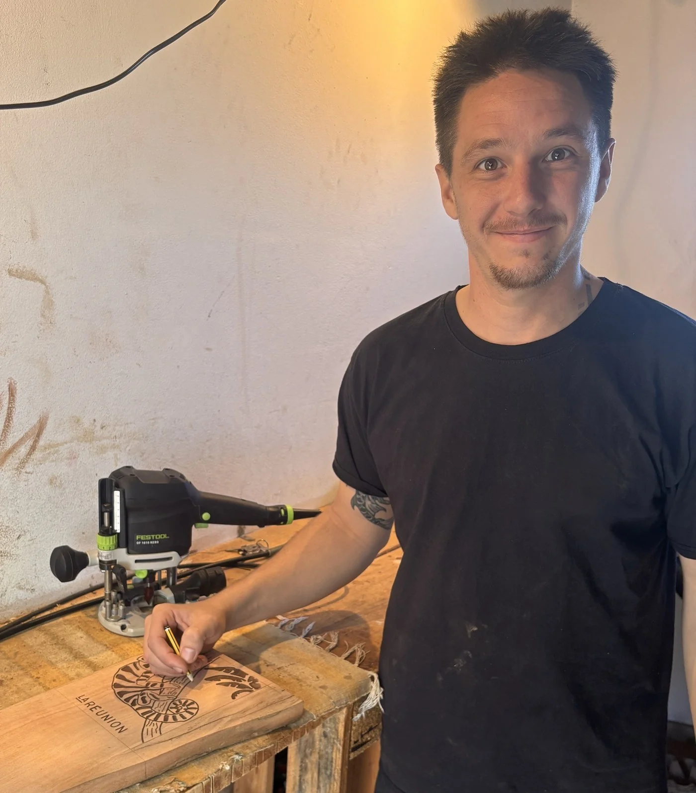 Tony Grandin à son établi d'Étang-Salé, dessinant un motif au crayon sur une planche, la défonceuse à côté